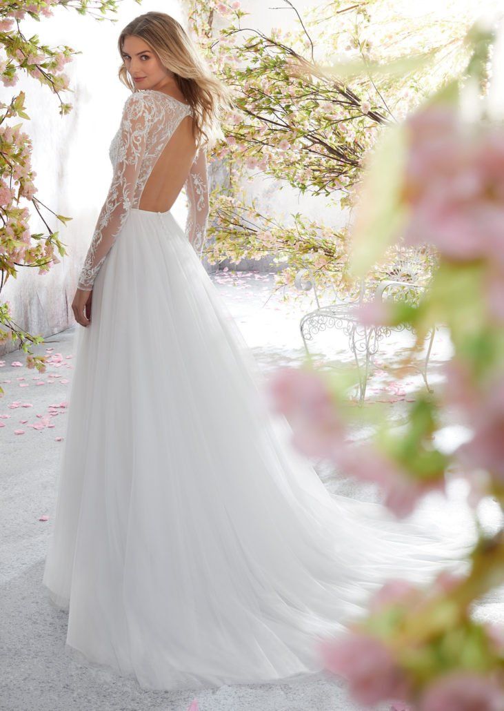Eine Frau trägt ein weißes Hochzeitskleid mit langen Ärmeln und tiefem Rückenausschnitt.