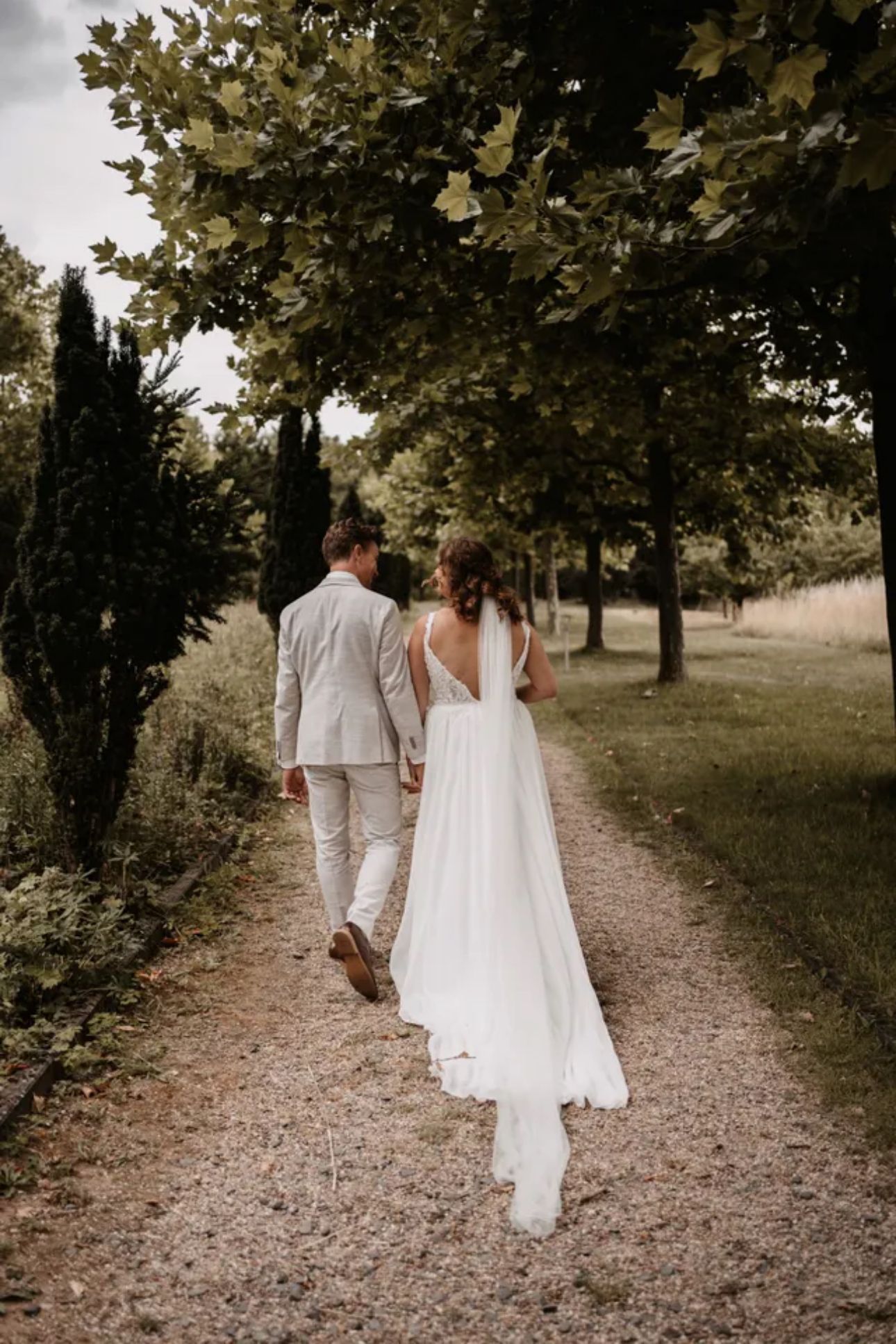 Paar in Hochzeitskleidung geht Hand in Hand einen von Bäumen umgebenen Weg entlang.
