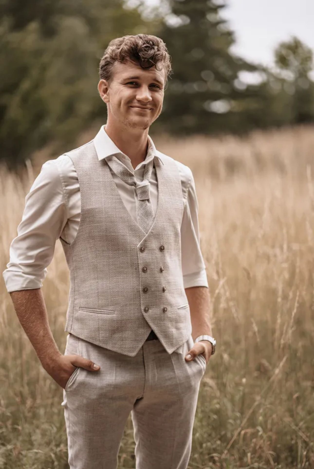 Junger Mann mit lockigem Haar lächelt, trägt Weste und Anzughose auf einem Feld.