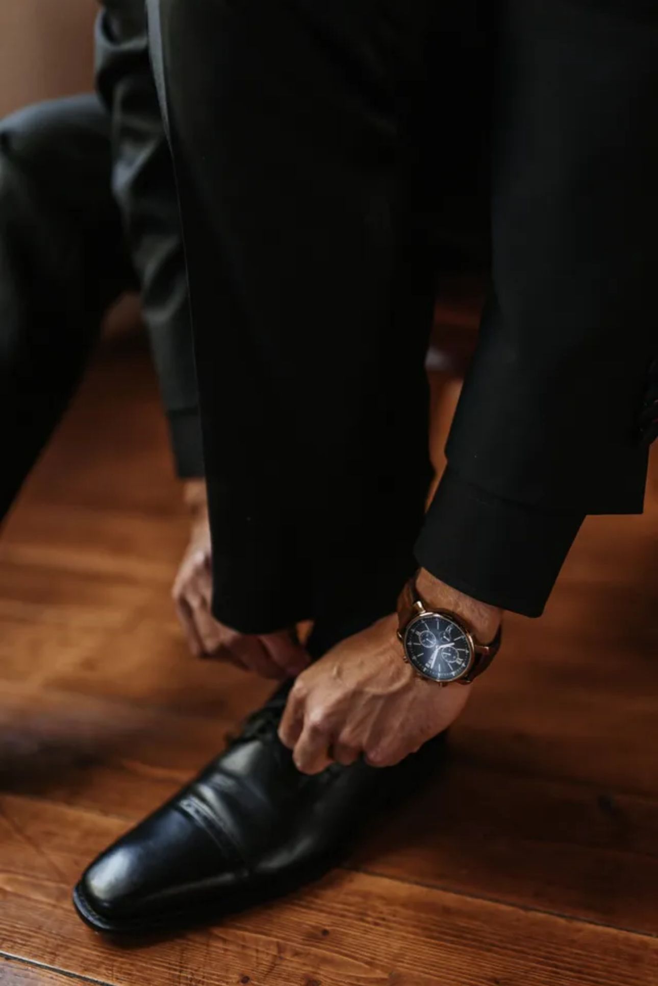 Mann im Anzug bindet schwarze Abendschuhe. Braune Lederuhr sichtbar. Holzboden.