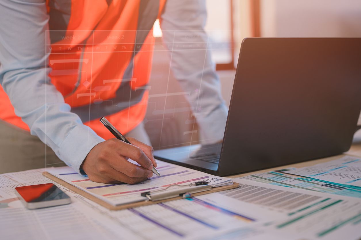A man wearing an orange safety vest over a light blue dress shirt signs a document next to an open laptop.