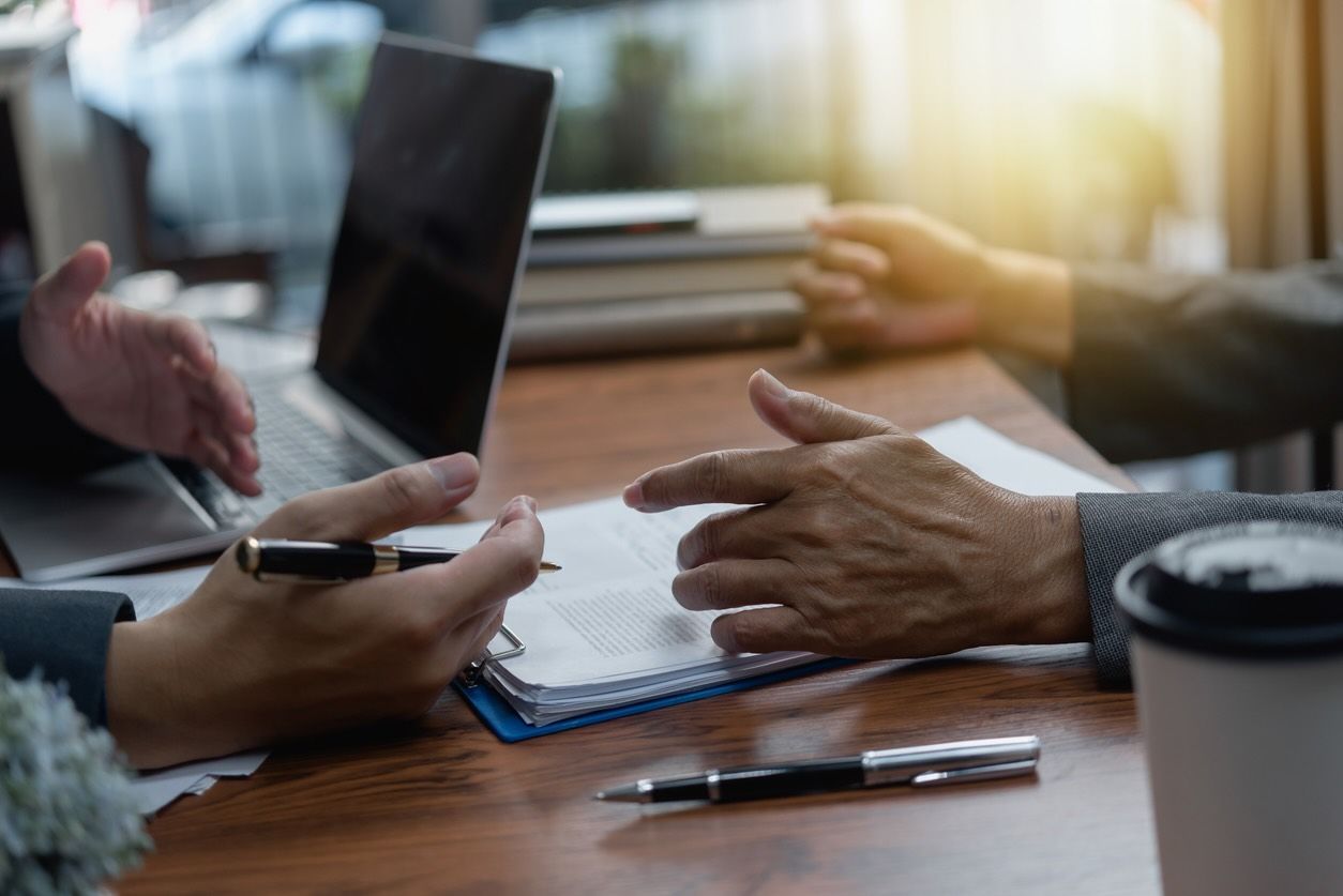Two people talk in a business environment at a table with an open laptop and a clipboard.