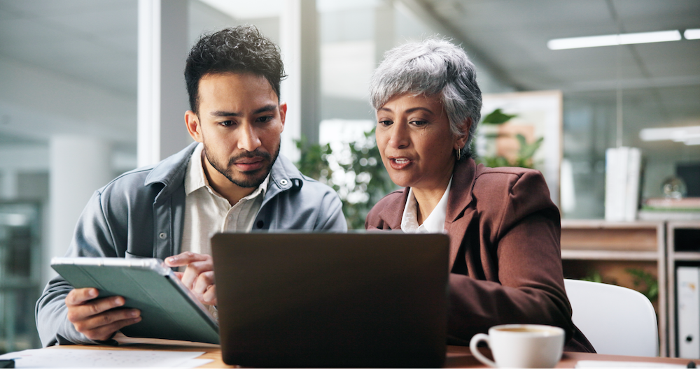 Man and woman in office space talking and working together on a laptop.