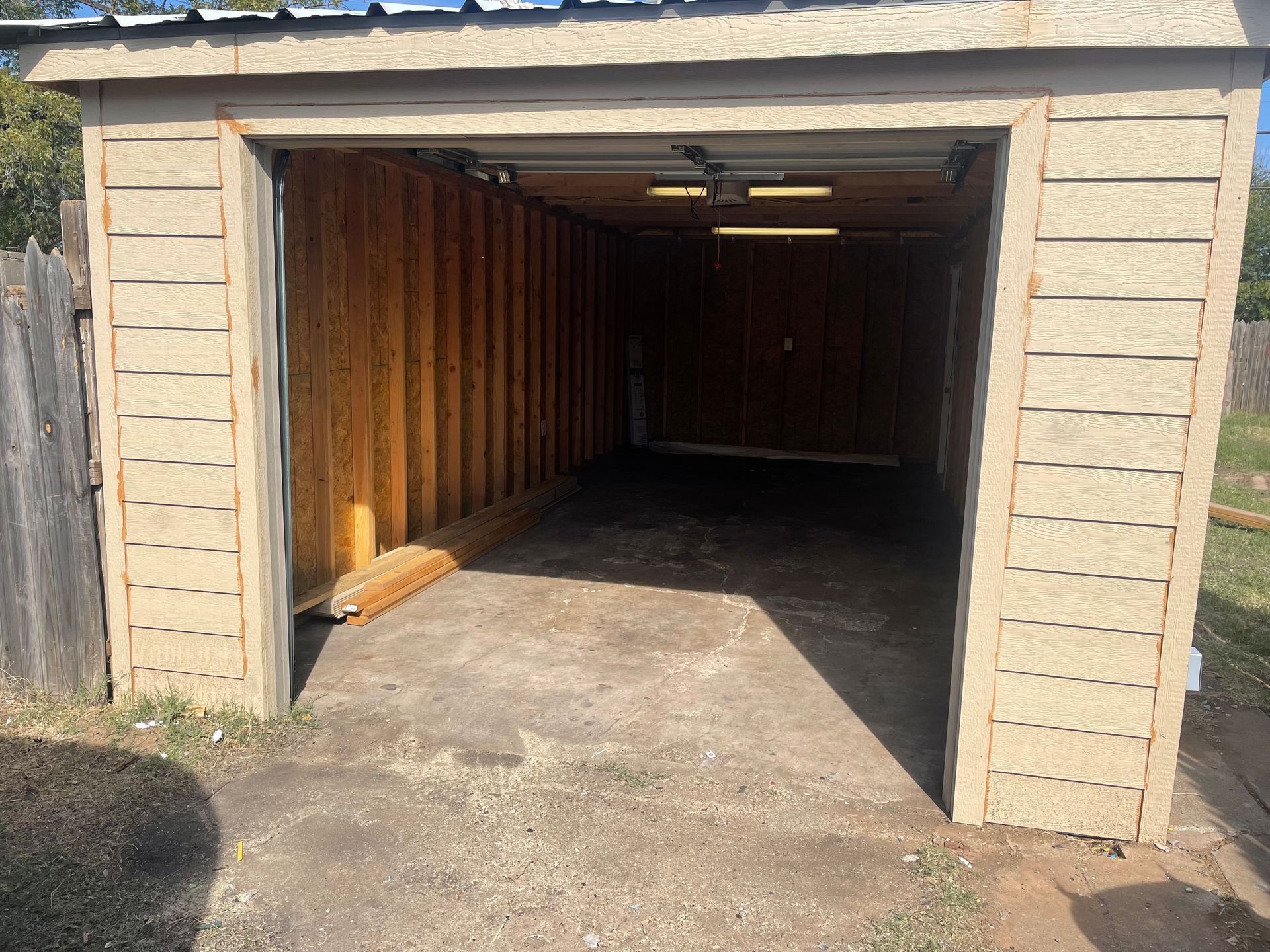 Open wooden garage with concrete floor, lit interior, and some boards leaning against the left wall