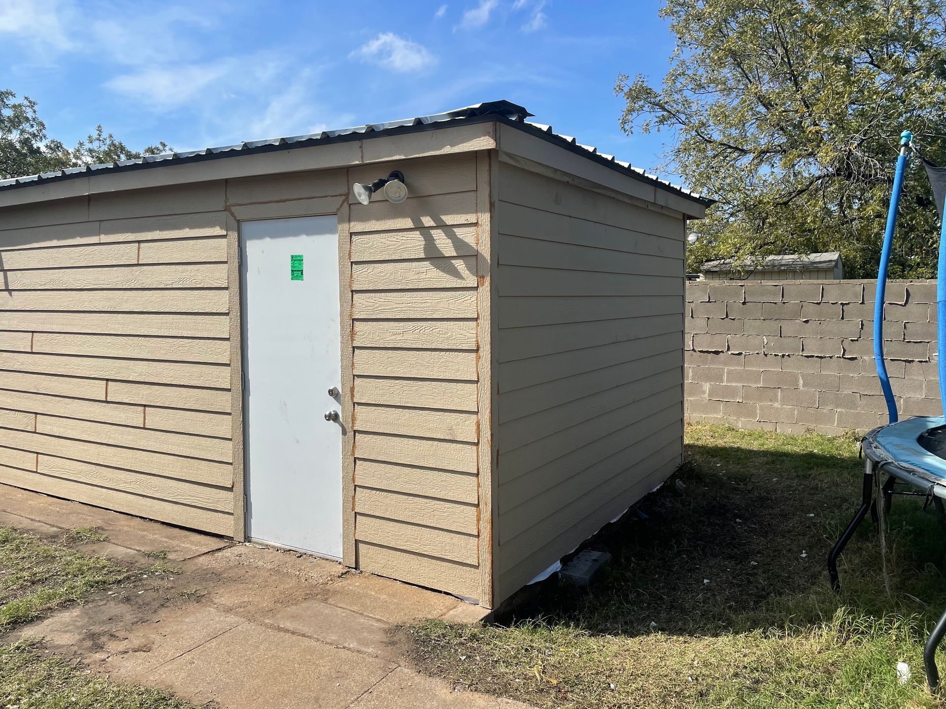 Small beige shed with a white door in a backyard, beside a trampoline and cinder block wall.
