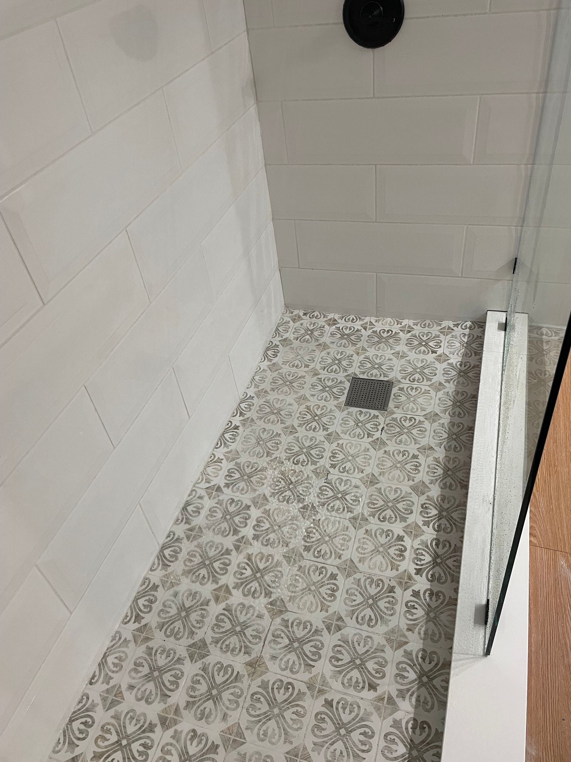 Empty shower stall with patterned floor tiles, gray wall tiles, and a drain near the back.
