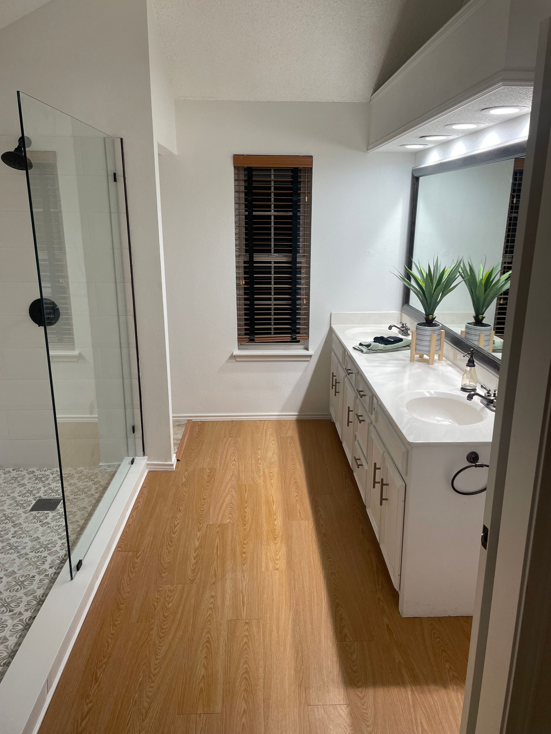 Bathroom with glass shower, white vanity, mirror, and wood-look floor.