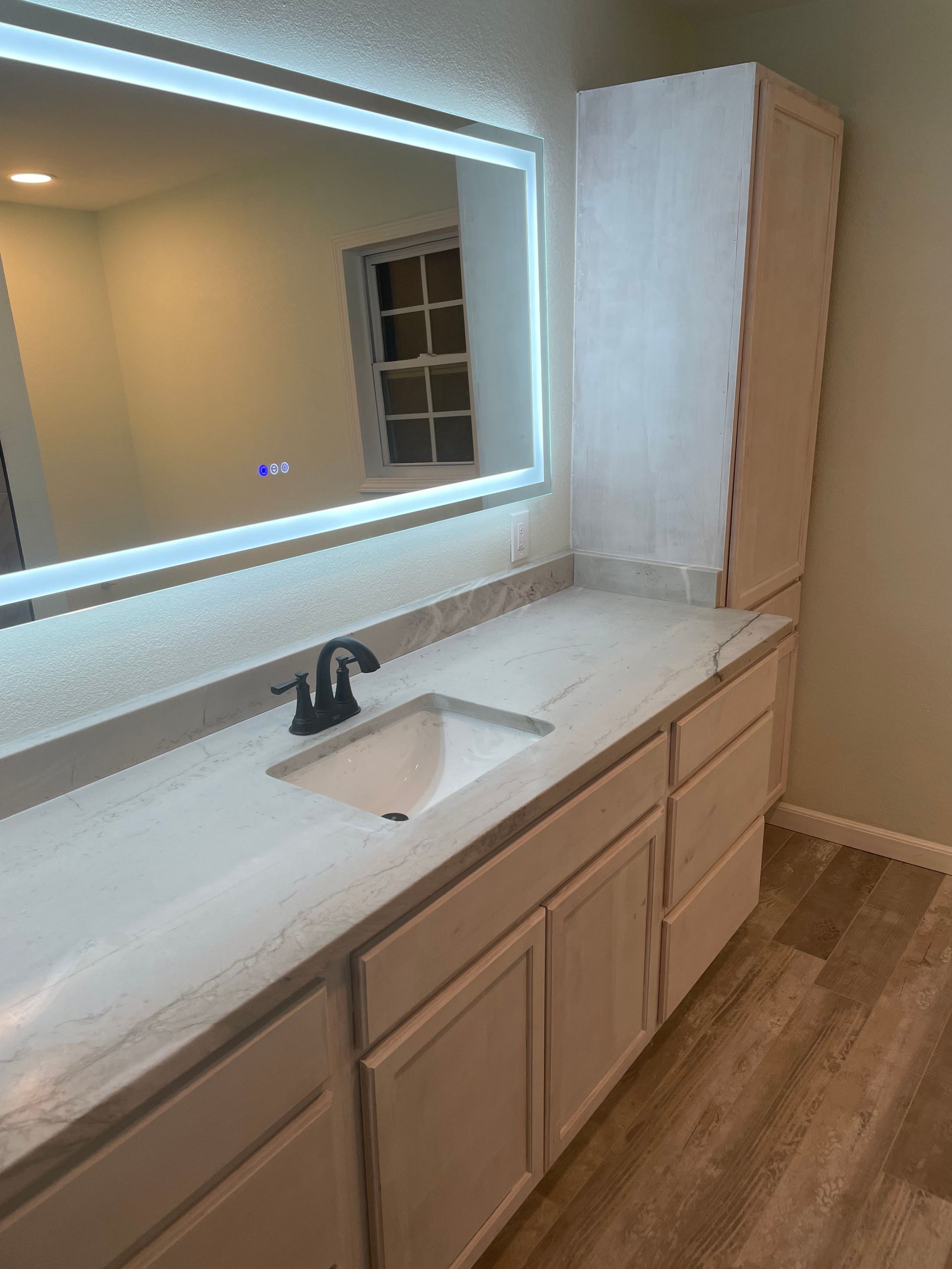 Bathroom vanity with white countertop, black faucet, large mirror, and tall cabinet, in a beige room.
