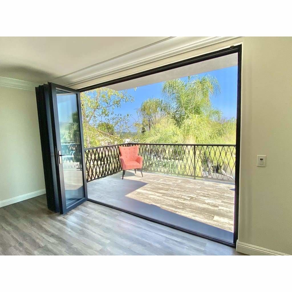 Open sliding glass doors to a sunny balcony with a red chair and leafy trees beyond