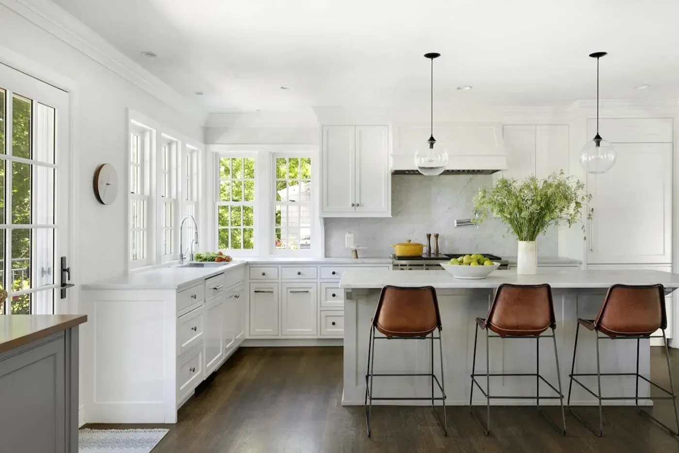 Bright white kitchen with island, leather stools, dark wood floors, and large windows under pendant lights