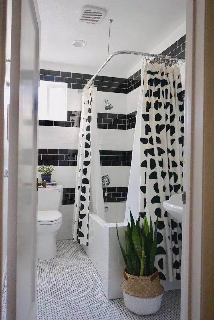 Narrow black-and-white tiled bathroom with shower curtain, toilet, sink, and potted plant.