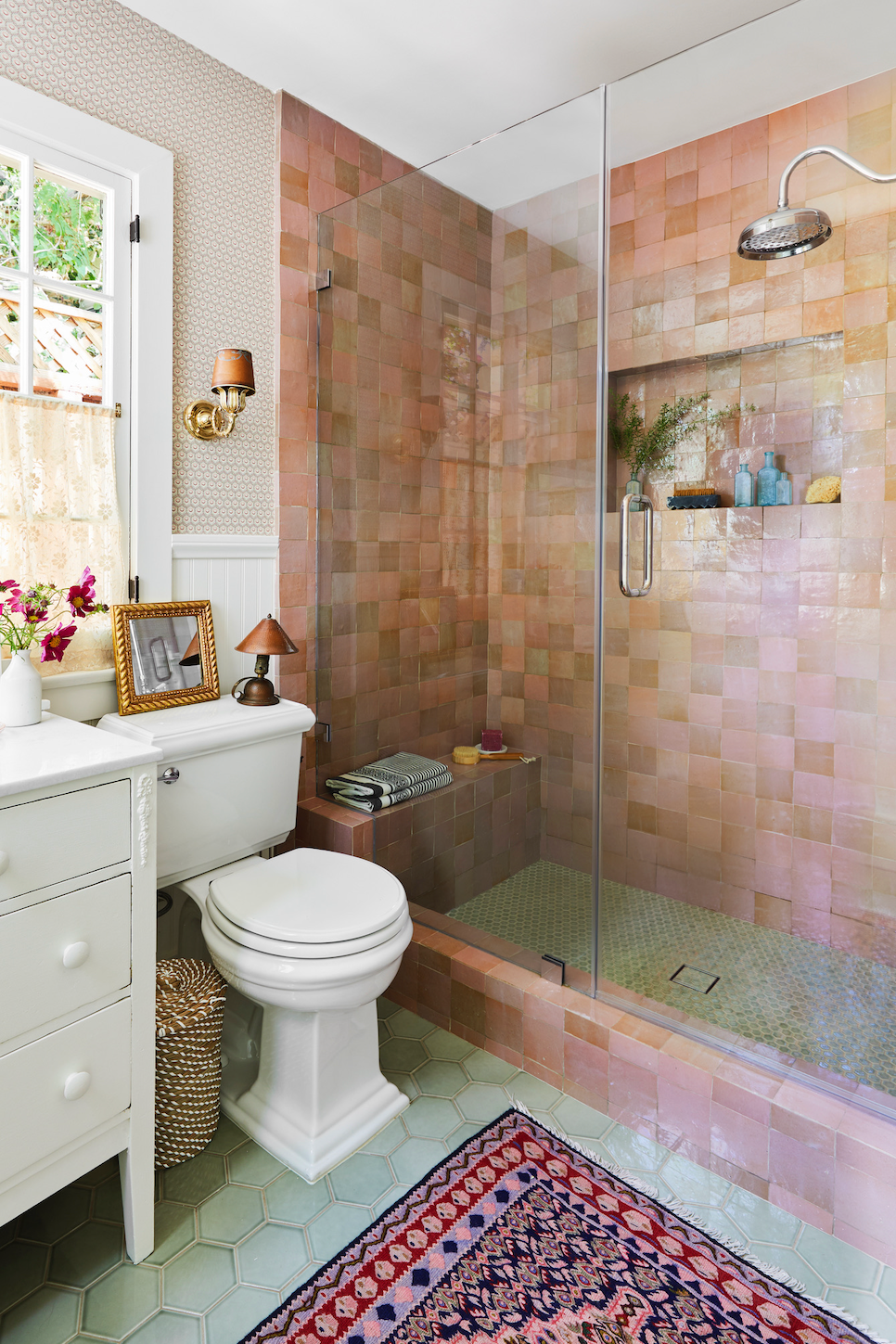 Compact bathroom with glass shower, white toilet, and vanity, in pink tile with a patterned rug
