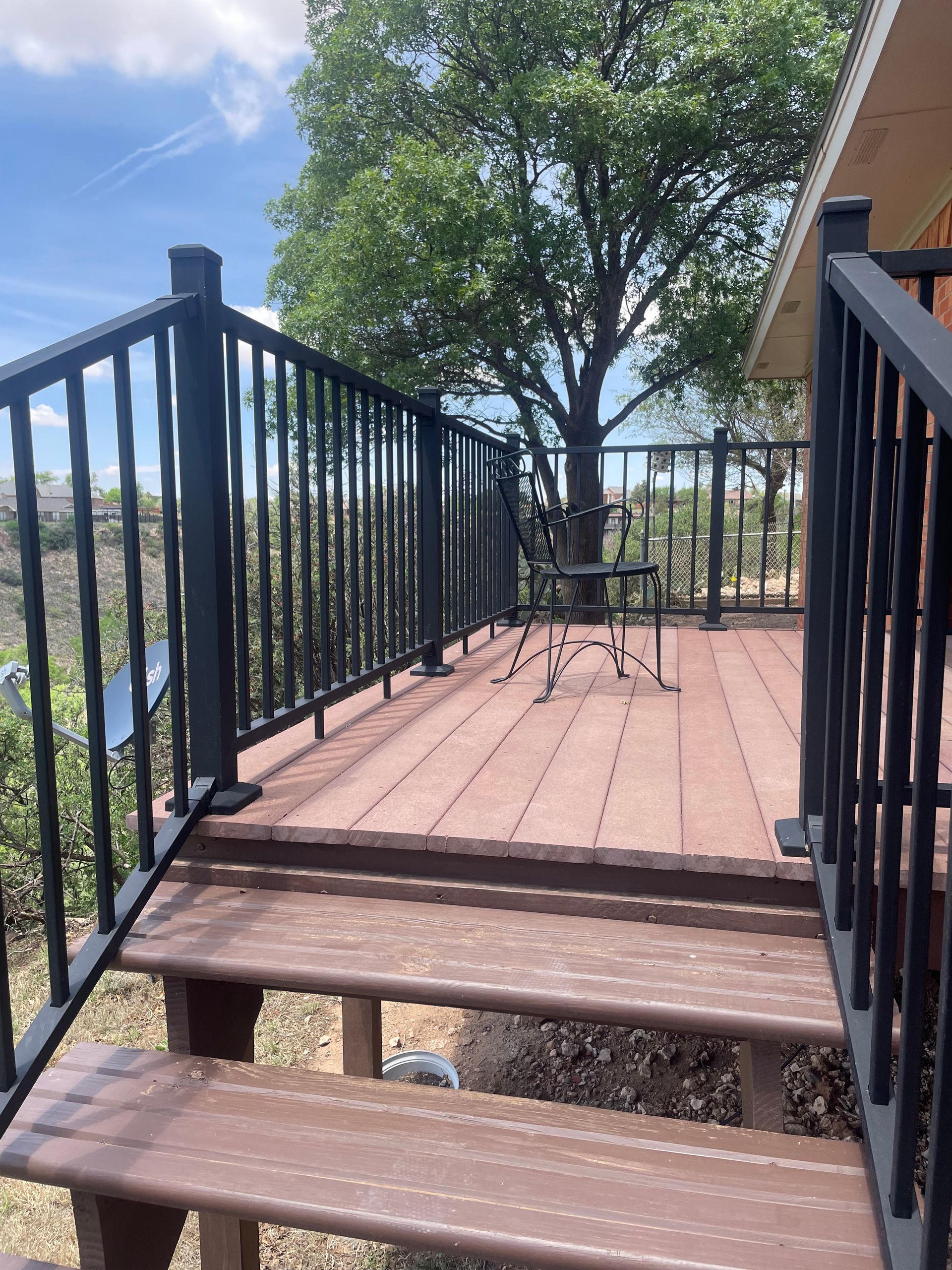 Wooden deck steps with black railings leading to a patio table and chairs under trees in daylight