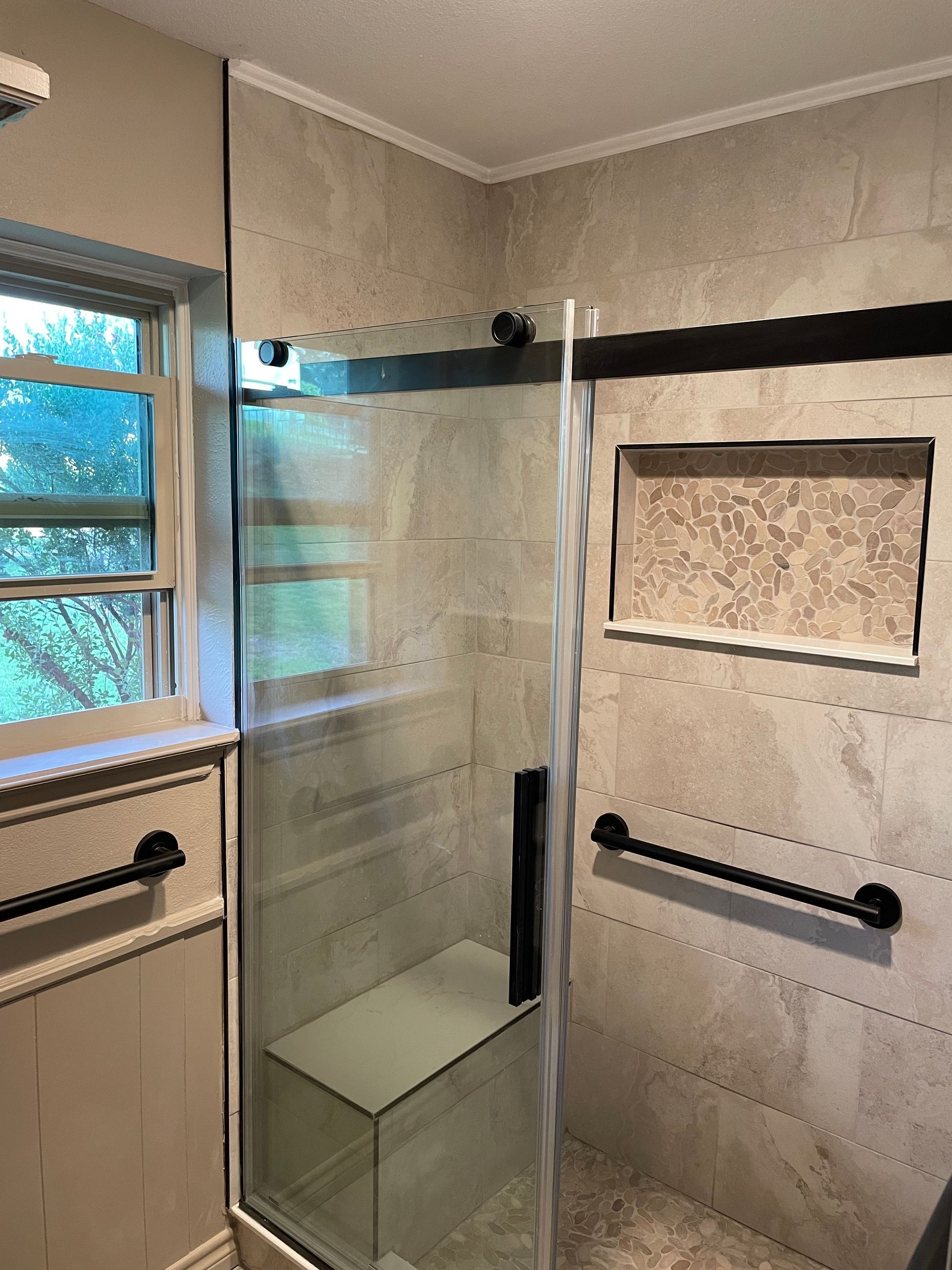 Modern bathroom with glass walk-in shower, black grab bars, and a bench beside a window.
