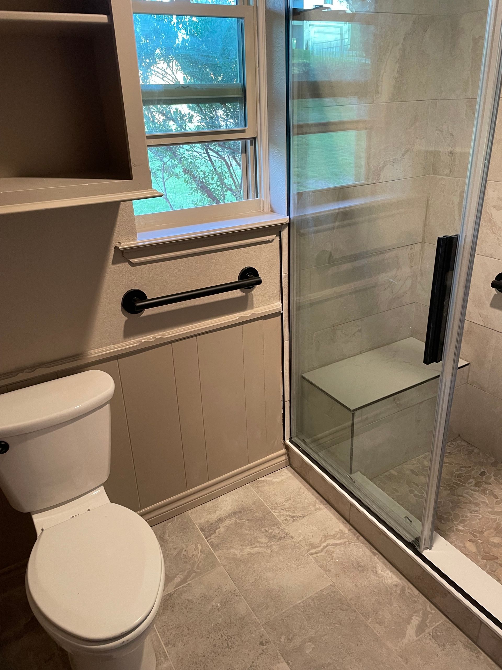 Small bathroom with toilet, window, grab bar, and glass shower enclosure.