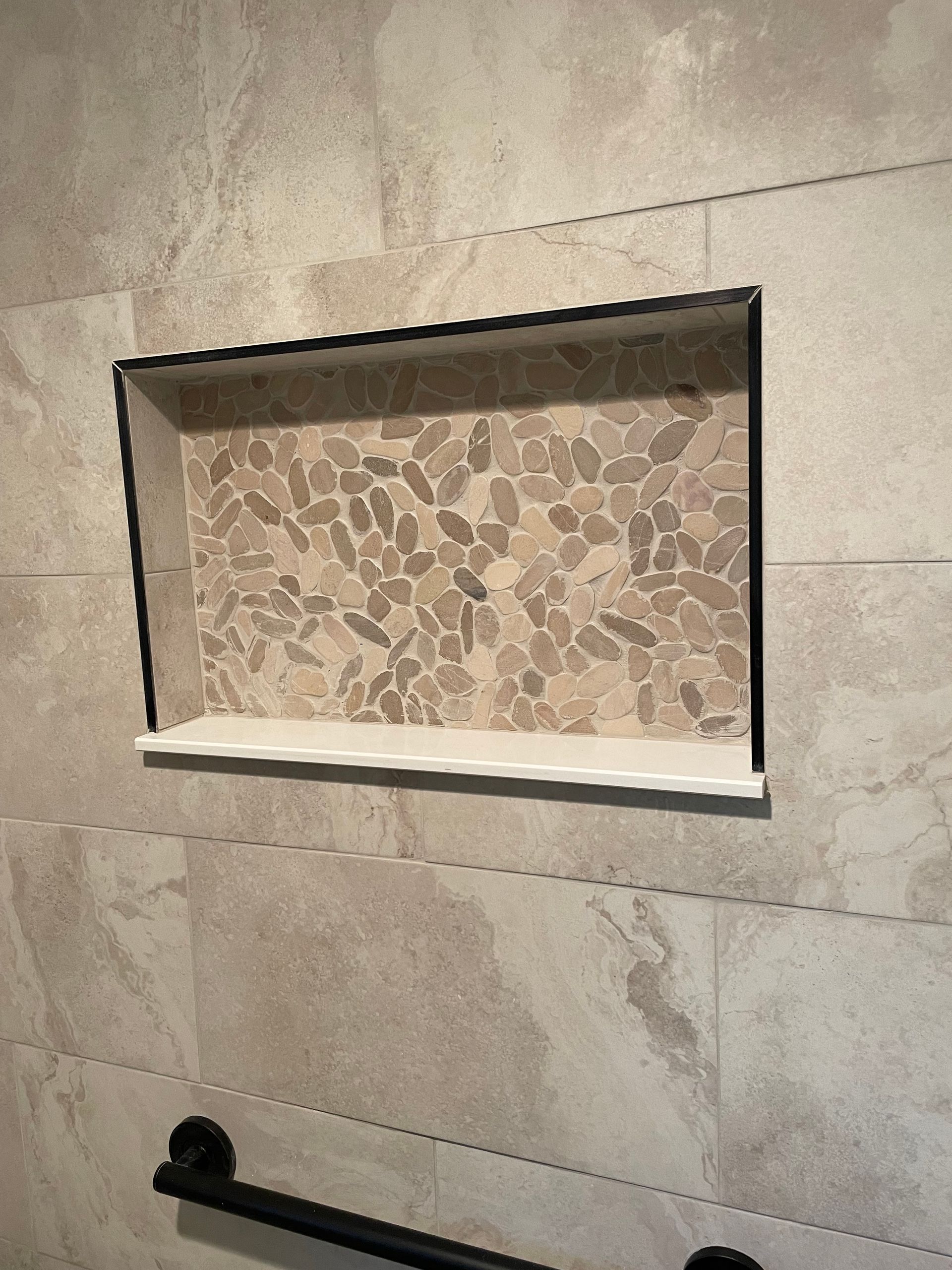 Empty shower niche with stone mosaic backing and a white shelf in a beige tiled bathroom