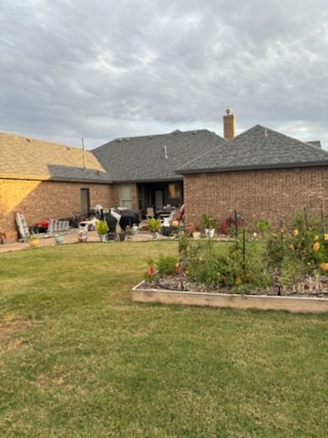 Backyard with brick house, lawn, flower beds, and two people near the patio under a cloudy sky