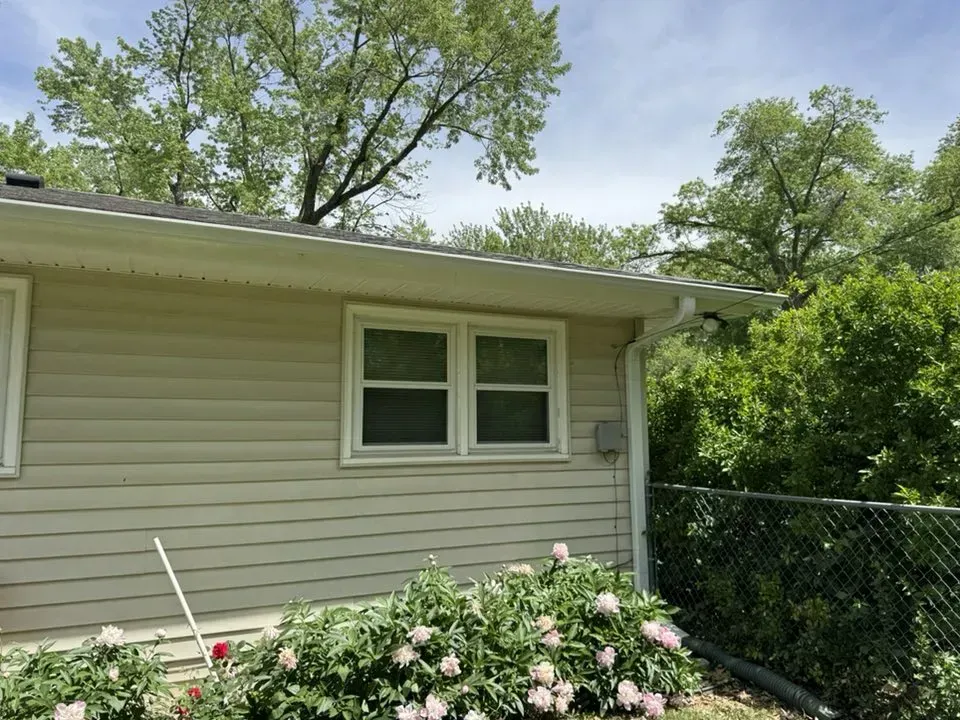 Tan house with white-framed windows, white gutters, and a chain-link fence, surrounded by green trees and bushes.