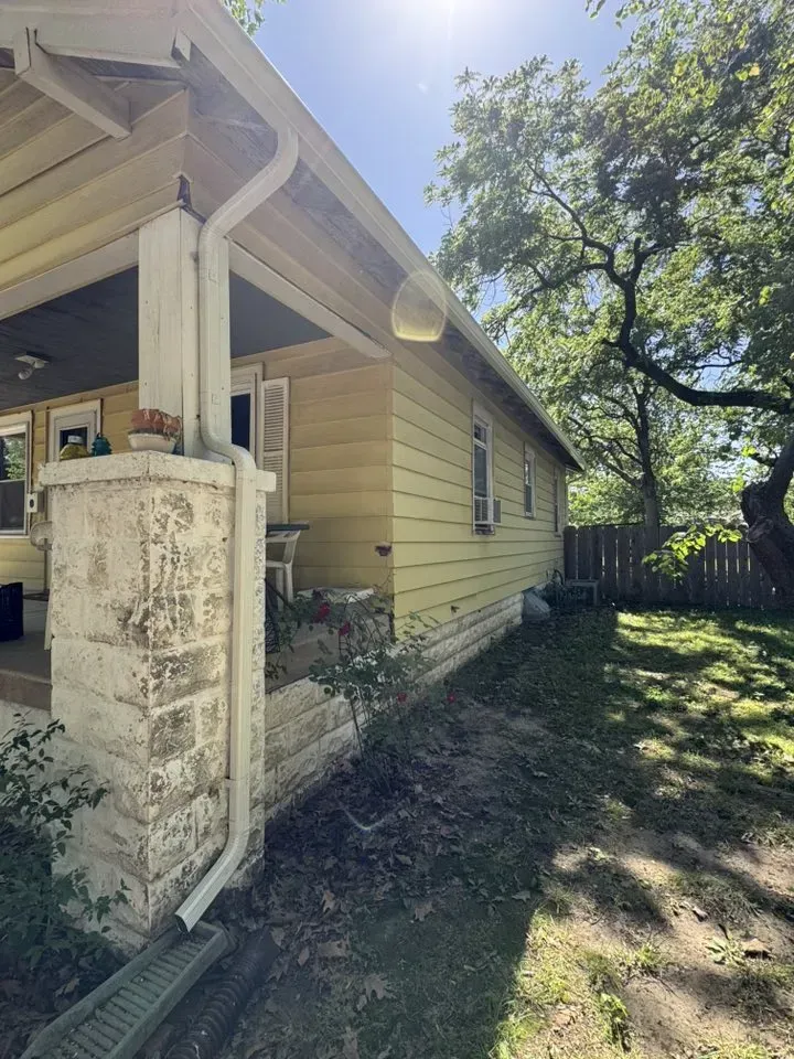 Yellow house exterior with porch, white trim and gutters newly replaced, and overgrown yard under a sunny sky.