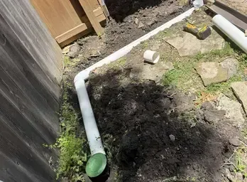 White PVC pipe drainage system being installed in a yard alongside a wooden fence.