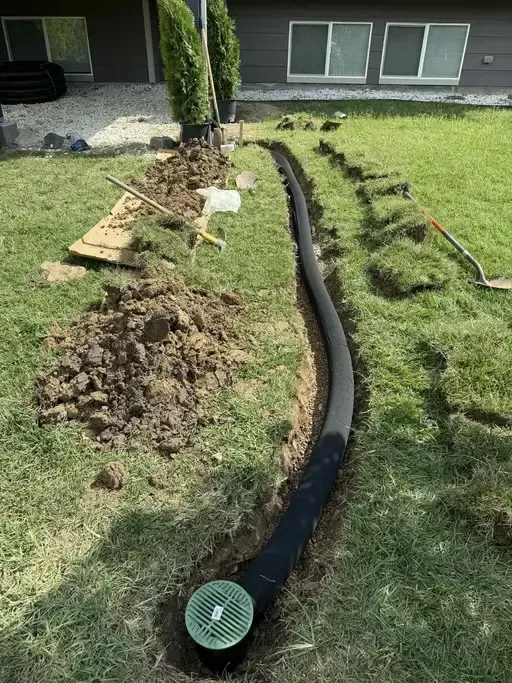 Before pic of a trench in a yard with a black drainage pipe, connected to a green grate, being installed.