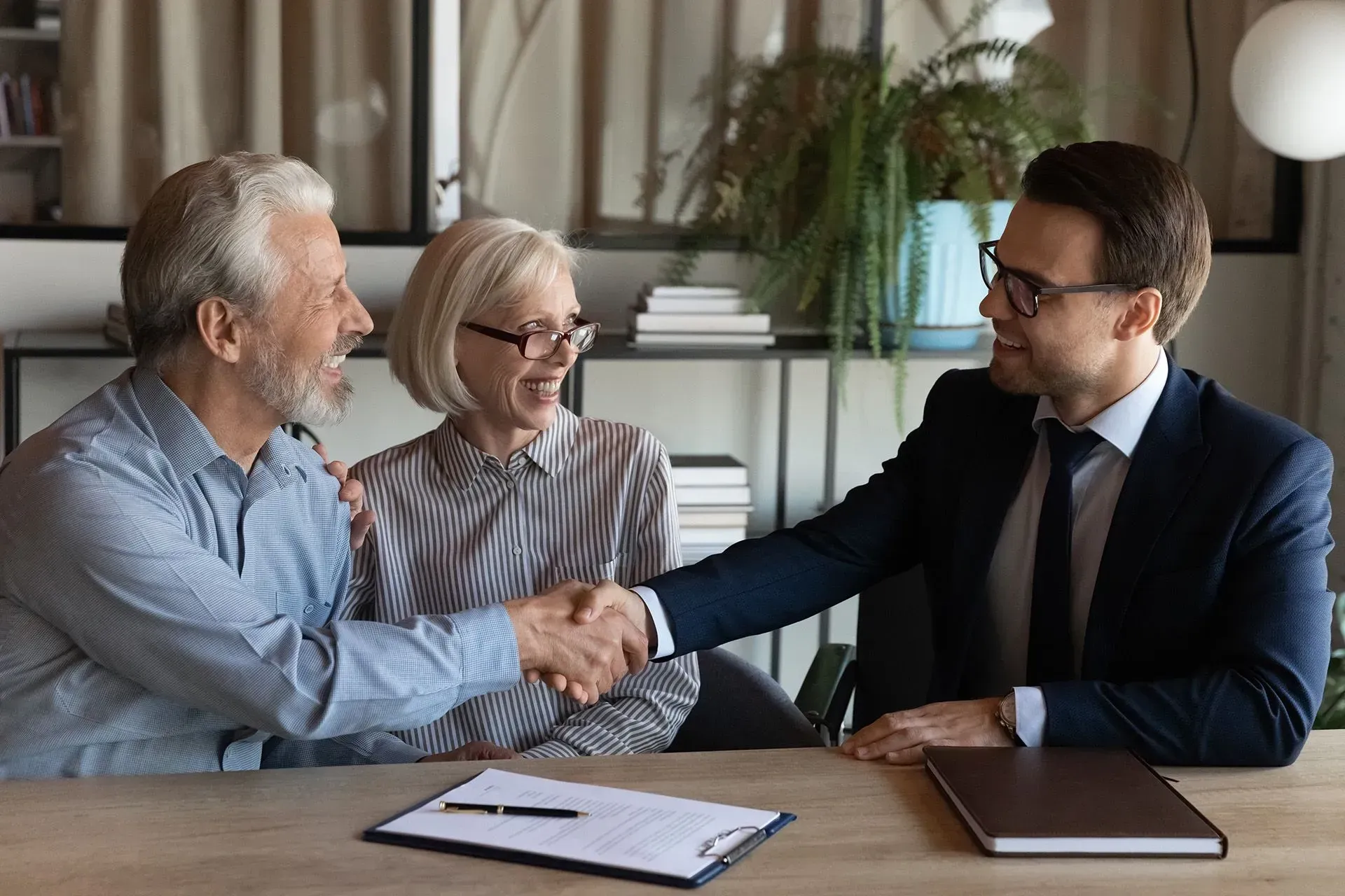 Two people shake hands across a table in a meeting room, with a third person smiling nearby.