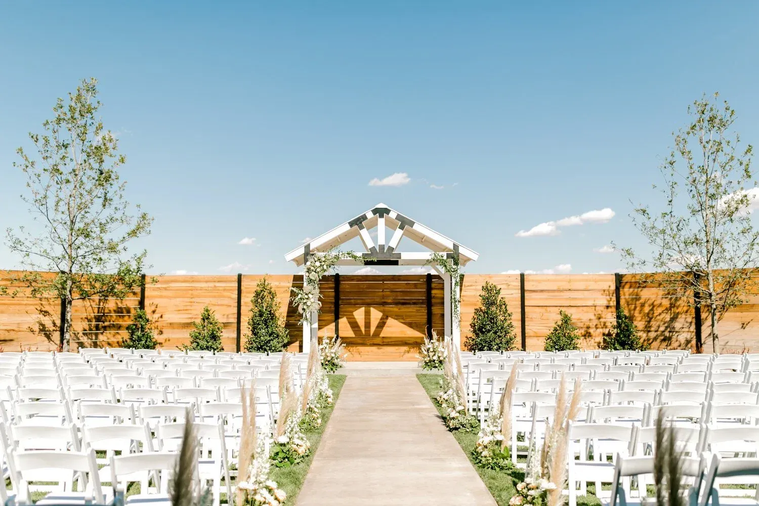 Outdoor wedding ceremony setup with white chairs, central aisle, and wooden arch under a clear blue sky