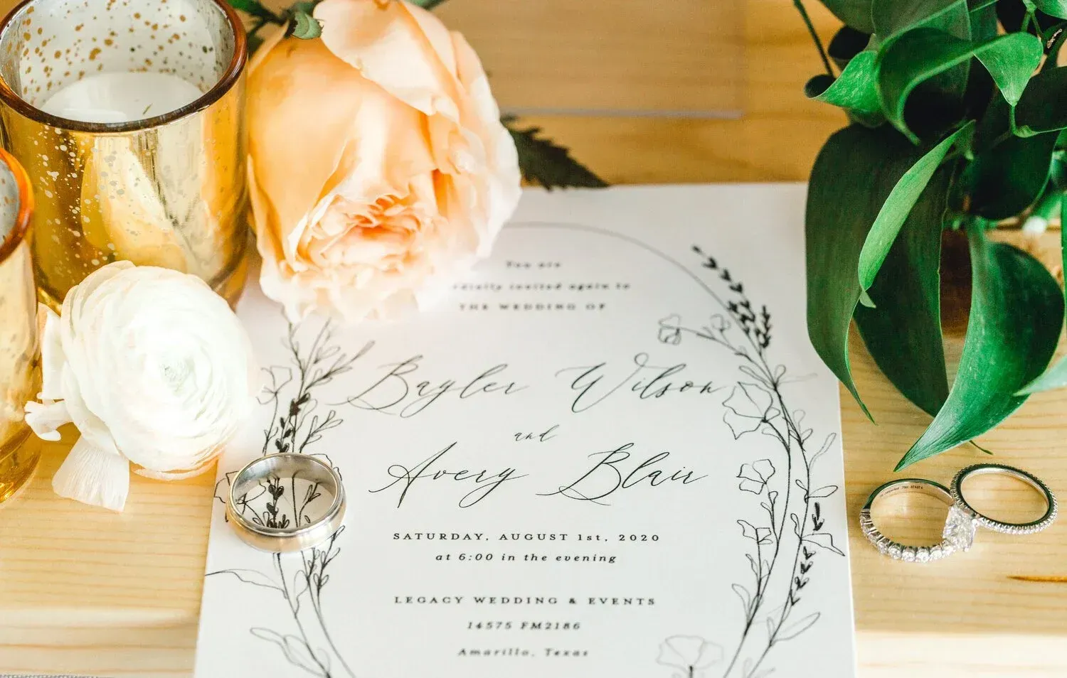 Wedding invitation with peach roses, greenery, a candle, and rings on a wooden table