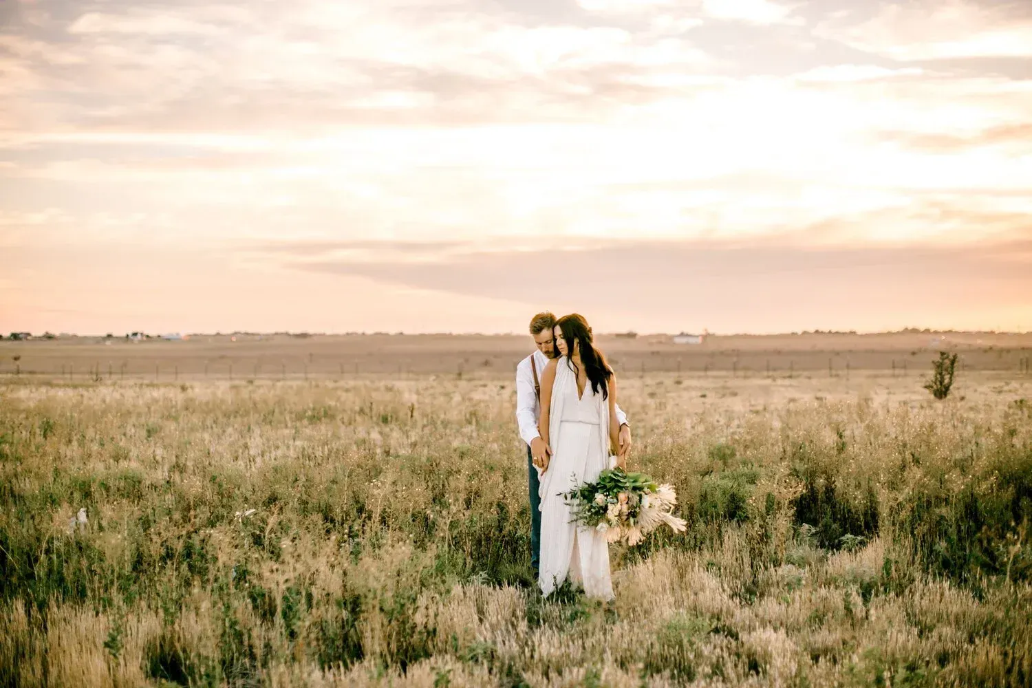 Two people in white standing in a grassy field at sunset, holding a bouquet