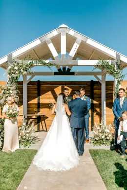 Bride and groom standing under a white wedding arbor outdoors, with floral arrangements and guests nearby