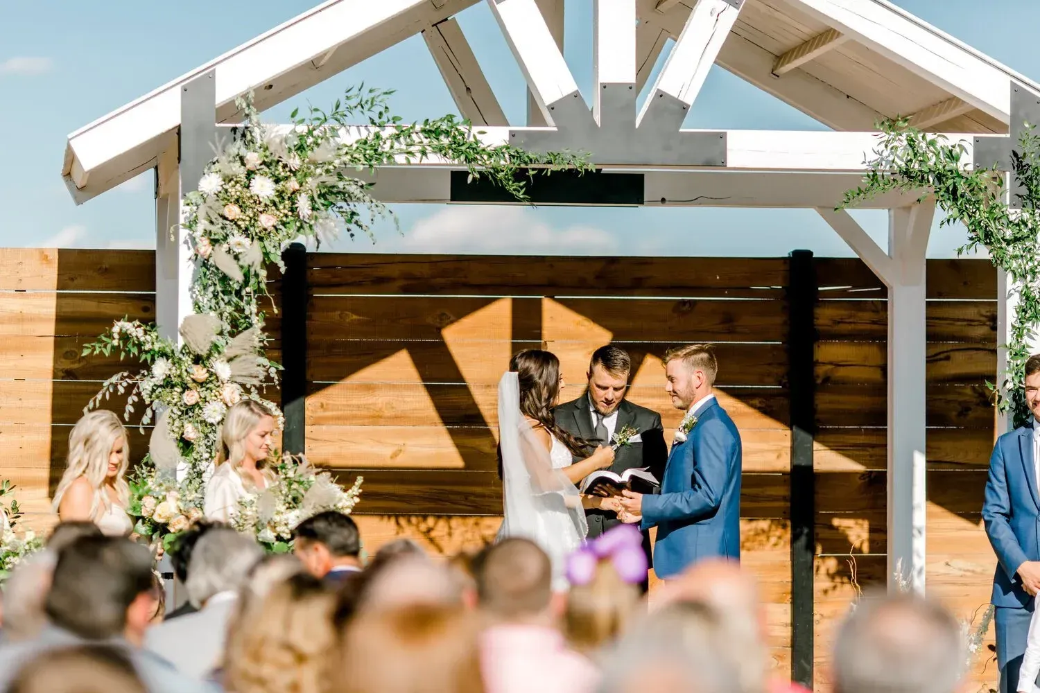 Wedding ceremony under a white floral arch, with a couple and officiant on a sunlit wooden stage.