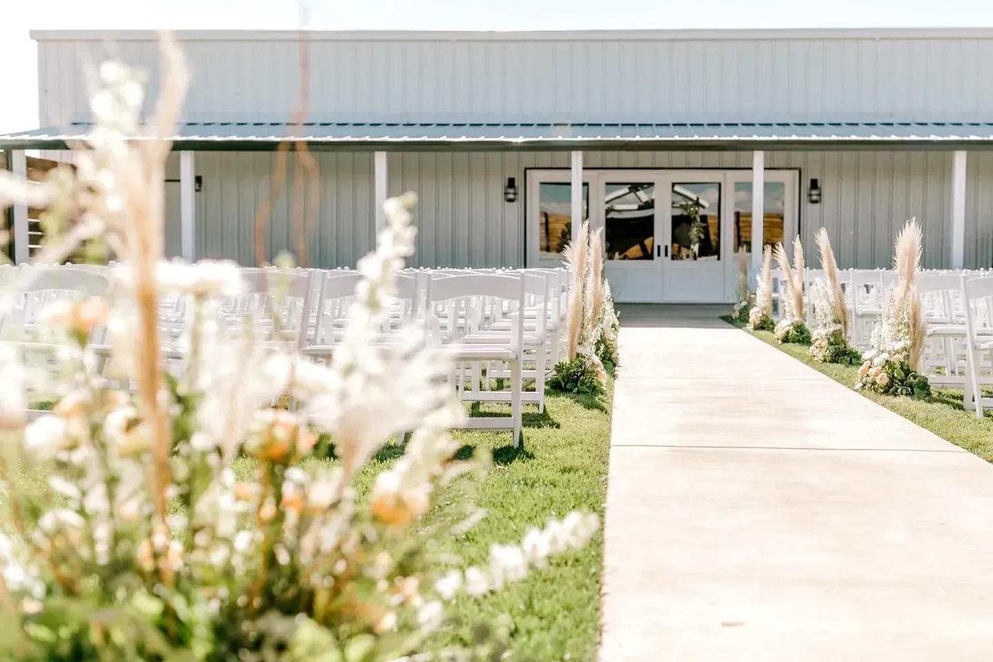 Outdoor wedding ceremony setup with white chairs, flower arrangements, and a central aisle leading to a barn entrance