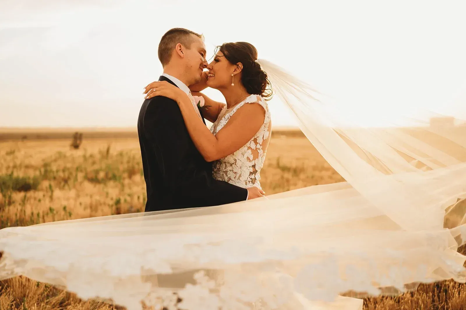 Couple embracing in a windy field, with a flowing veil at sunset
