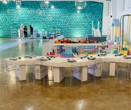 Colorful art studio with a long white table, chairs, and a turquoise wall of craft supplies