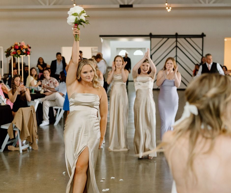 Bridal party walking down aisle, bride in satin dress holding bouquet overhead at indoor wedding reception