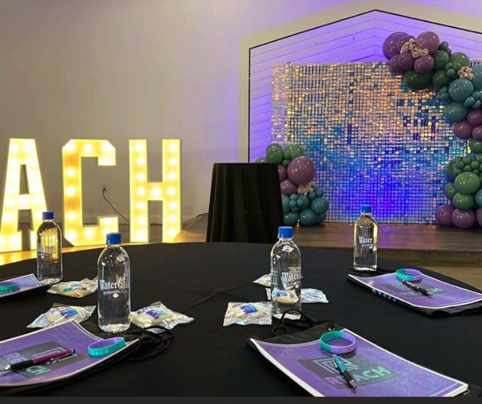 Decorated party table with water bottles, black-and-purple settings, and lit “ACH” letters beside a balloon arch