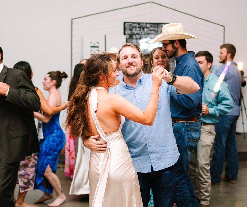 People dancing at a lively indoor party, with a woman in a white dress and a man in a light blue shirt in front.