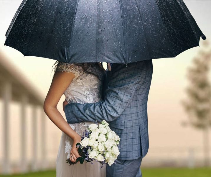 Couple embracing under a black umbrella, with the bride holding a white bouquet in a rainy outdoor setting.