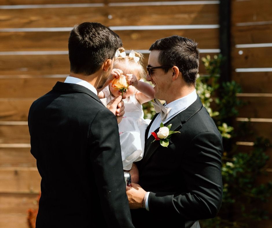 Two men in suits holding a small child outdoors in front of a wooden fence.