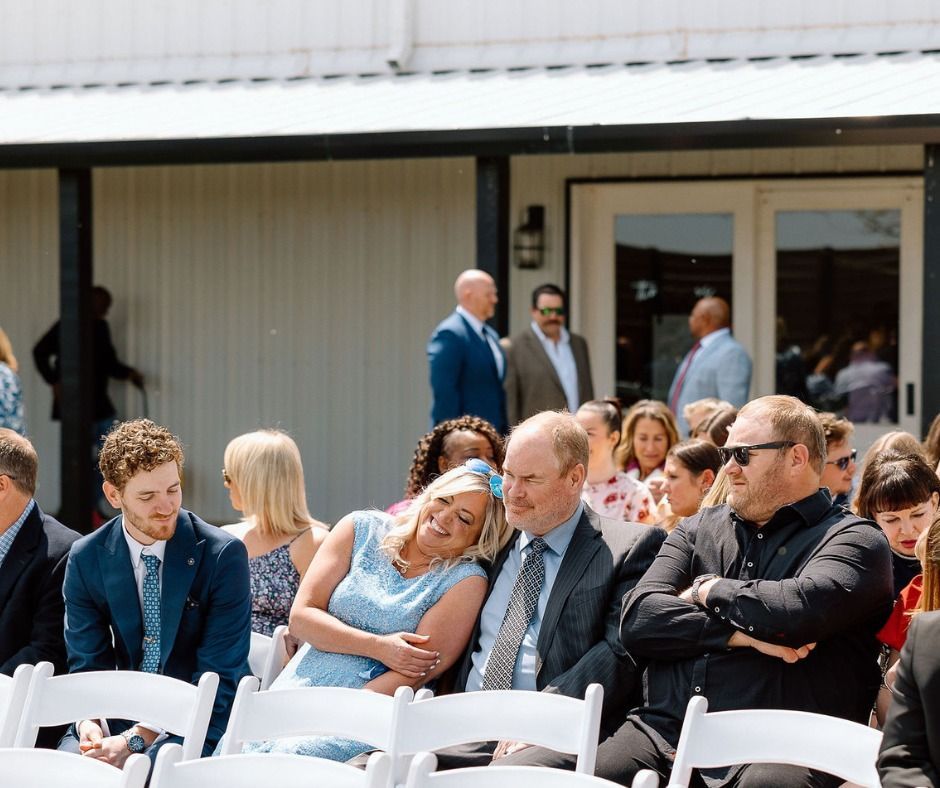 Seated audience in white chairs at an outdoor event, chatting and waiting near a building