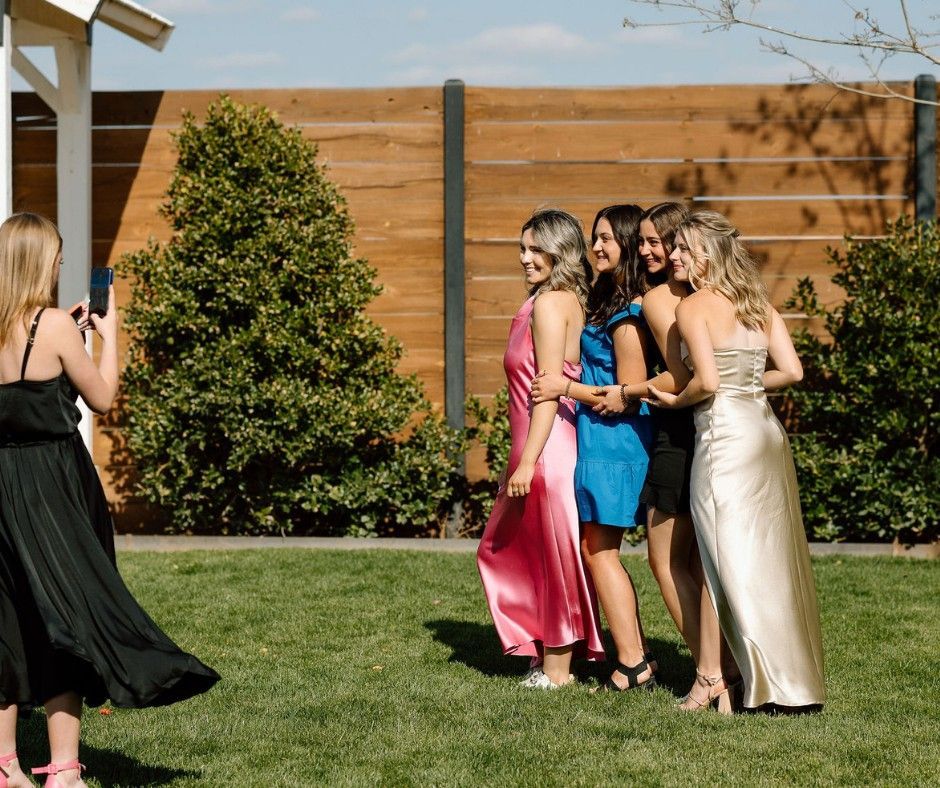 Four women posing outdoors on a sunny lawn, with a wooden fence and shrubs behind them.