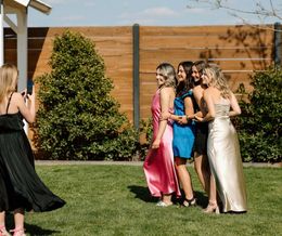 Four women in formal dresses posing and smiling on a lawn outdoors, with a photographer at left.