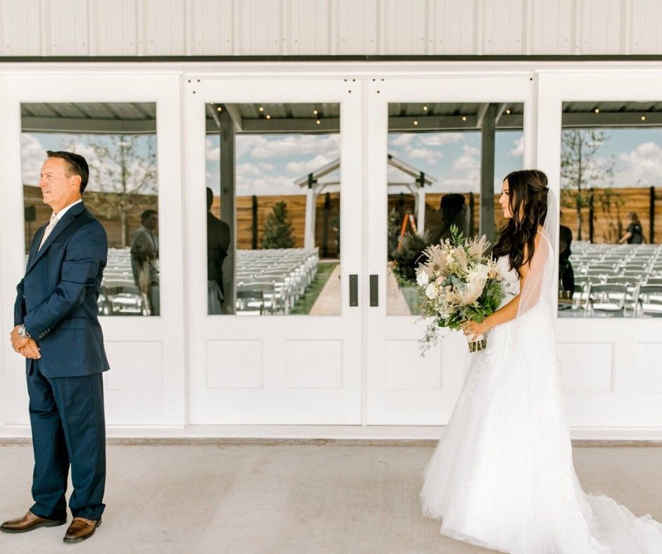 Bride and groom stand apart in front of white wedding venue doors, holding a bouquet.
