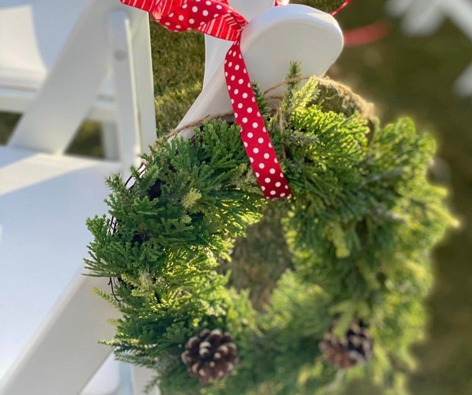 Small evergreen wreath with pinecones and a red polka-dot ribbon hanging on a white chair outdoors