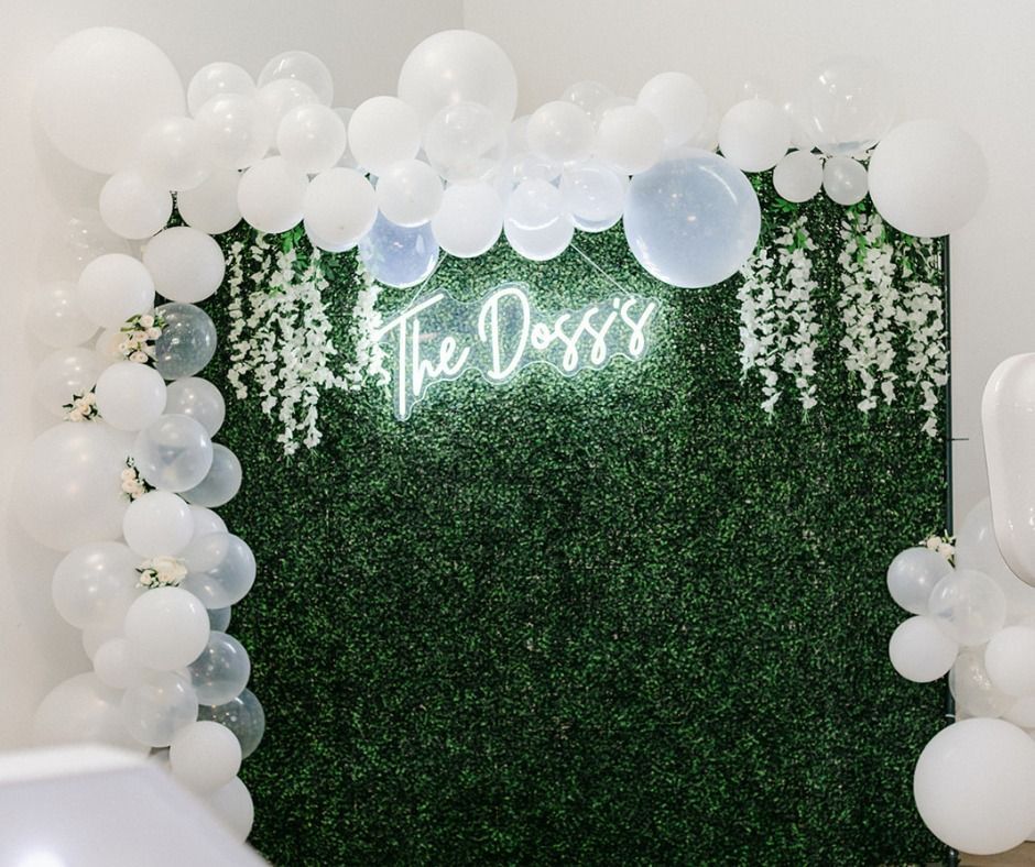 Green floral backdrop with white balloon garland and neon sign reading “The Doss”