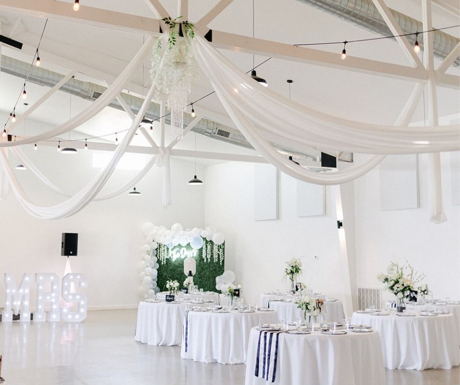 Bright white wedding reception hall with draped ceiling fabric, round tables, floral arrangements, and “MR & MRS” lights