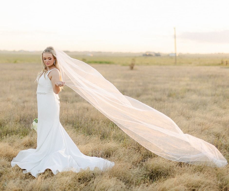Woman in a white gown standing in a grassy field, her long veil trailing in the wind at sunset.