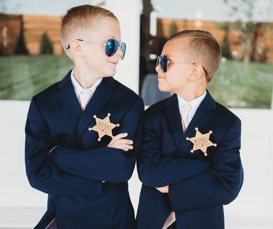 Two children in navy suits and sunglasses stand with arms crossed, wearing star-shaped badges, outdoors.