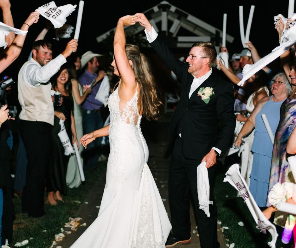 Newlyweds raise their arms while guests cheer with white napkins at an outdoor wedding reception