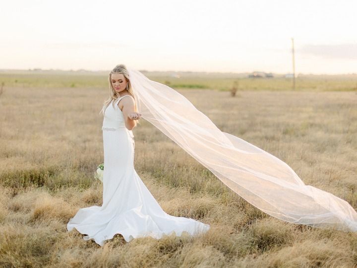 Woman in a white dress stands in a grassy field with a long veil flowing behind her.
